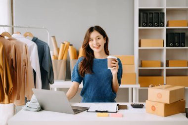 Asyalı terzi kadın terzi atölyesinde giysiler üzerinde çalışıyor. Stüdyoda dizüstü bilgisayarı, telefonu ve kahvesi olan güzel bir bayan moda tasarımcısı.
