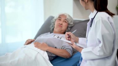 Beyaz takım elbiseli Asyalı doktor hastanede yatan Asyalı yaşlı kadın hastayı muayene ederken not alıyor.
