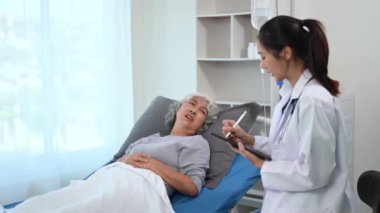 Beyaz takım elbiseli Asyalı doktor hastanede yatan Asyalı yaşlı kadın hastayı muayene ederken not alıyor.