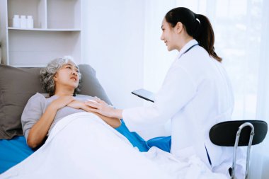 Beyaz takım elbiseli Asyalı doktor hastanede yatan Asyalı yaşlı kadın hastayı muayene ederken not alıyor.