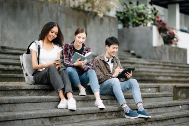 Genç üniversite öğrencileri parktaki merdivenlerde tablet, akıllı telefon ve kitap okuyarak ders çalışıyor. 