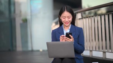 Şehir caddesinde cep telefonu ve dizüstü bilgisayarla çalışan mutlu Asyalı iş kadını.