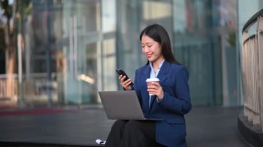 Şehir caddesinde kahve içerken cep telefonu ve dizüstü bilgisayarla çalışan mutlu Asyalı iş kadını.