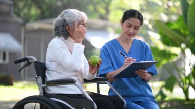 Asyalı doktor hastane parkında tekerlekli sandalyedeki yaşlı bir kadını muayene ederken not alıyor.