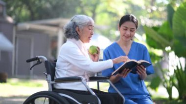 Genç Asyalı hemşire, hastane parkında tekerlekli sandalyedeki yaşlı kadına kitap okuyor. Kıdemli sağlık hizmeti kavramı