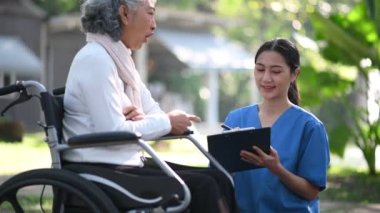 Asyalı doktor hastane parkında tekerlekli sandalyedeki yaşlı bir kadını muayene ederken not alıyor.
