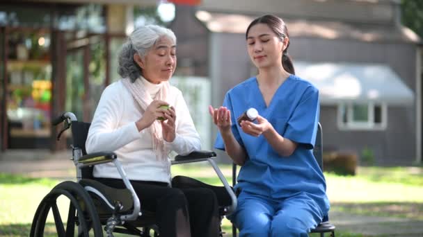 若いアジアの看護師が病院の公園の車椅子の上のシニア女性に錠剤を与えています シニアヘルスケアコンセプト — ストック動画