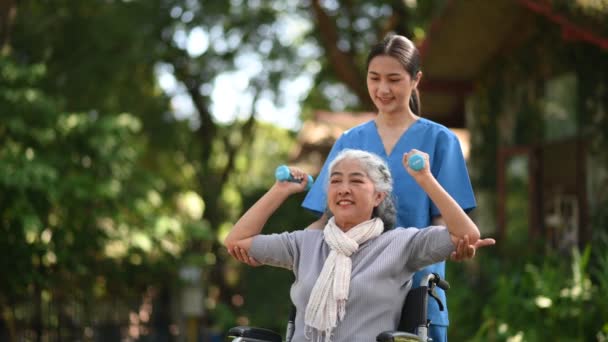 アジアの若い女性医師は 病院の公園でダンベルで運動するために車椅子の上のシニア女性を助けます シニアヘルスケアコンセプト — ストック動画