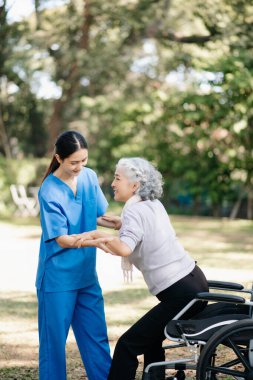 Asyalı genç bakıcı hemşire dışarıda yürüyen yaşlı bayana destek oluyor. Uzman doktor yaşlı yetişkinlere yardım edip onlarla ilgilenir. Huzurevi hastane bahçesi konsepti