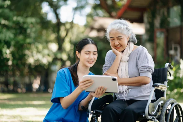 stock image Elderly asian senior woman on wheelchair with Asian careful caregiver and encourage patient using digital tablet  in garden