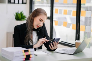 Akıllı telefondan konuşan genç ve güzel bir kadın çalışma yerinde otururken hesap makinesi ve dizüstü bilgisayar kullanıyor. Modern ofiste işe odaklanmış.