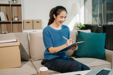 Genç Asyalı kadın evdeki kanepede dinleniyor ve panoya yazı yazarken dizüstü bilgisayar kullanıyor. 