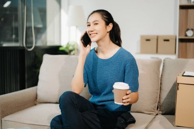 Genç Asyalı kadın evdeki kanepede dinleniyor ve akıllı telefonla konuşuyor ve kahve içiyor.