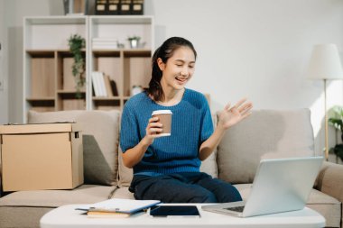 Genç Asyalı kadın evdeki kanepede dinleniyor ve kahve içerken dizüstü bilgisayar kullanıyor.   