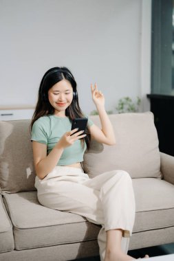 Genç Asyalı kadın evinde rahat bir kanepede rahat rahat akıllı telefondan mesaj atıyor.