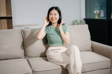 Genç Asyalı kadın evdeki rahat kanepeye uzanıp kulaklık kullanıyor. 