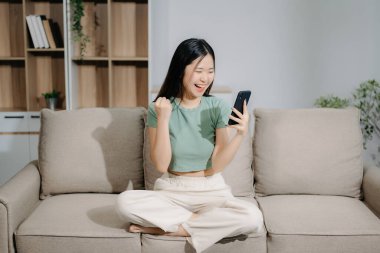 Genç Asyalı kadın evdeki kanepede otururken akıllı telefon kullanıyor.