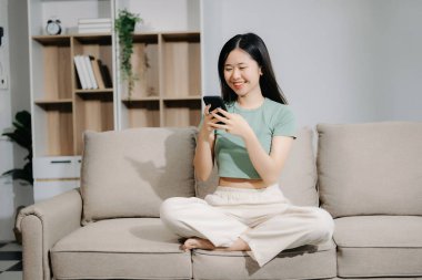 Genç Asyalı kadın evdeki kanepede otururken akıllı telefon kullanıyor.