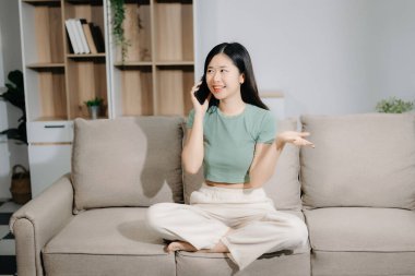 Genç Asyalı kadın evdeki kanepede otururken akıllı telefon kullanıyor.
