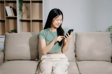 Asyalı genç bir kadın evdeki kanepede akıllı telefon kullanıyor.