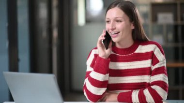 Genç ve güzel bir iş kadını, modern ofisteki masada otururken bilgisayarla çalışıyor ve akıllı telefonla konuşuyor.