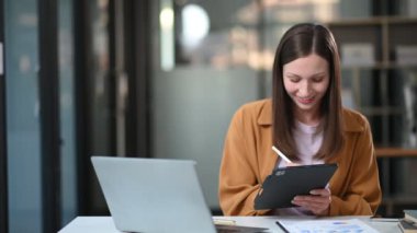 Modern ofisteki masada otururken dijital tablet ve laptop üzerinde çalışan genç ve güzel bir iş kadını.