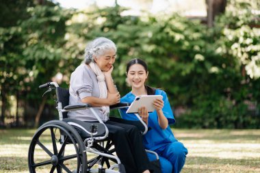 Tekerlekli sandalyedeki yaşlı Asyalı yaşlı kadın ve bahçede dikkatli bir bakıcı.