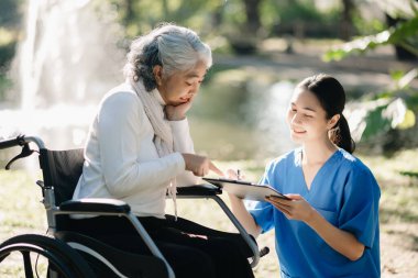 Tekerlekli sandalyedeki yaşlı Asyalı yaşlı kadın ve bahçede dikkatli bir bakıcı.