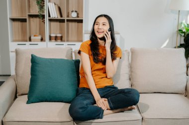 Mutlu Asyalı kadın evdeki kanepede akıllı telefon kullanıyor.