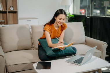 Genç Asyalı kadın rahat koltuğunda oturup not alıyor ve dizüstü bilgisayar kullanıyor.