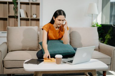 Asyalı kadın ev ofisteki kanepede dizüstü bilgisayarla çalışmaktan yoruldu ve çok düşünüyor.