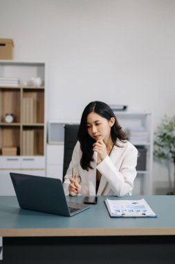 Modern ofisteki masada akıllı telefon ve dizüstü bilgisayar kullanan Asyalı güzel bir iş kadını.