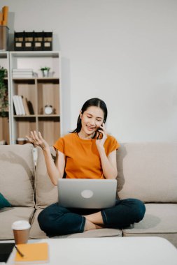 Genç Asyalı kadın evdeki kanepede dinleniyor ve akıllı telefonla konuşuyor, dizüstü bilgisayar kullanıyor.  