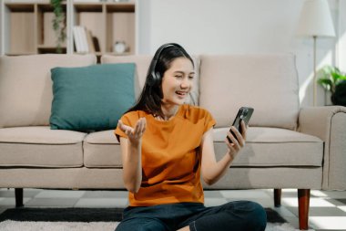 Evdeki kanepede otururken akıllı telefon ve kulaklık kullanan mutlu Asyalı kadın. 