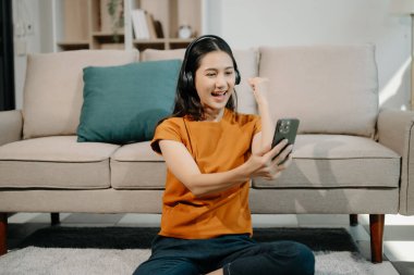 Evdeki kanepede otururken akıllı telefon ve kulaklık kullanan mutlu Asyalı kadın. 