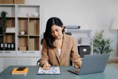Modern ofisteki masada akıllı telefon ve dizüstü bilgisayar kullanan Asyalı güzel bir iş kadını.