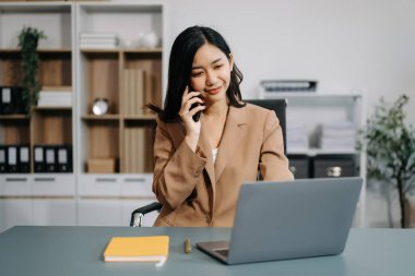 Modern ofisteki masada akıllı telefon ve dizüstü bilgisayar kullanan Asyalı güzel bir iş kadını.
