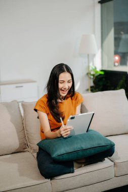 Genç Asyalı kadın evdeki rahat koltukta dijital tablet kullanarak rahatlıyor. 