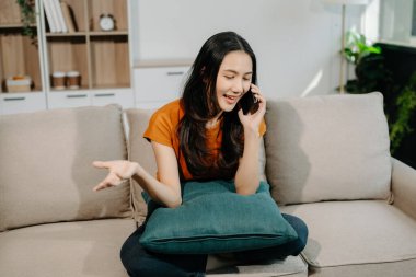 Genç Asyalı kadın evdeki rahat koltukta akıllı telefon kullanarak rahatlıyor.