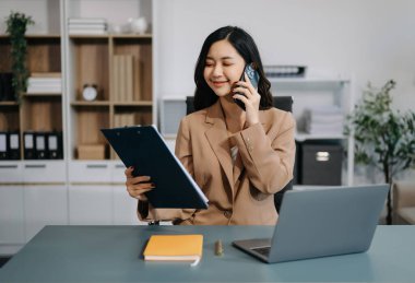 Genç ve güzel bir kadın akıllı telefondan konuşuyor ve çalışma yerinde otururken belgelere bakıyor. Modern ofiste işe odaklanmış.