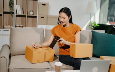 Başlangıçta küçük işletme KOBİ 'si, akıllı telefon ve laptop kullanan girişimci kadın, paket ürün kutularını hazırlamak için çevrimiçi alışveriş siparişi alıyor ve kontrol ediyor