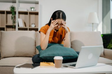 Asyalı kadın ev ofisteki kanepede dizüstü bilgisayarla çalışmaktan yoruldu ve çok düşünüyor.