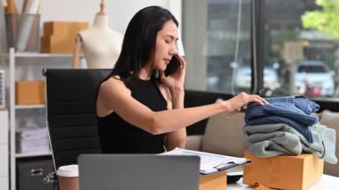  Moda tasarımcısının masasındaki Asyalı genç kadın stüdyoda çalışıyor, dizüstü bilgisayar kullanıyor ve akıllı telefondan konuşuyor. 