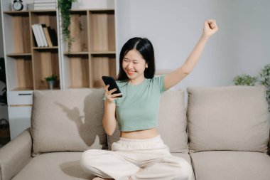 Mutlu Asyalı kadın evdeki koltukta otururken akıllı telefon kullanıyor.