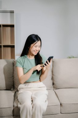 Asyalı genç bir kadın evdeki kanepede akıllı telefon kullanıyor.