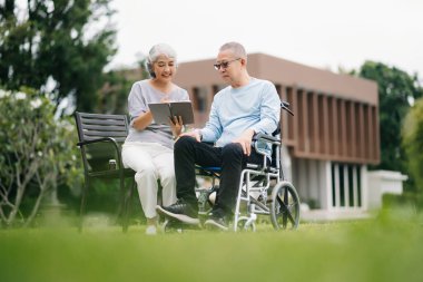 Asyalı yaşlı çift ve tekerlekli sandalyeli defter parkta birbirlerine göz kulak oluyorlar.