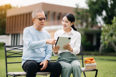 Asyalı yaşlı, üst düzey bir adam. Asyalı, dikkatli bir bakıcı. Tablet tableti ve hastane bahçesi konseptinde yiyecek..