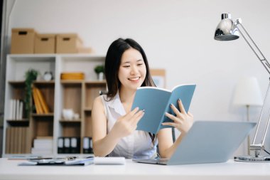 Genç, çekici Asyalı kadın ofis çalışanı gülümsemesi, elinde not defteri ve modern ofiste dizüstü bilgisayar kullanımı.