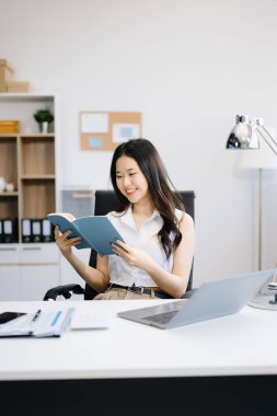 Genç Asyalı kadın ofiste otururken kitap okuyor.