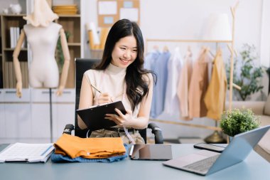 Asyalı terzi kadın terzi atölyesinde giysiler üzerinde çalışıyor. Modern stüdyoda başarılı olduktan sonra güzel genç bayan moda tasarımcısı gülümsemesi